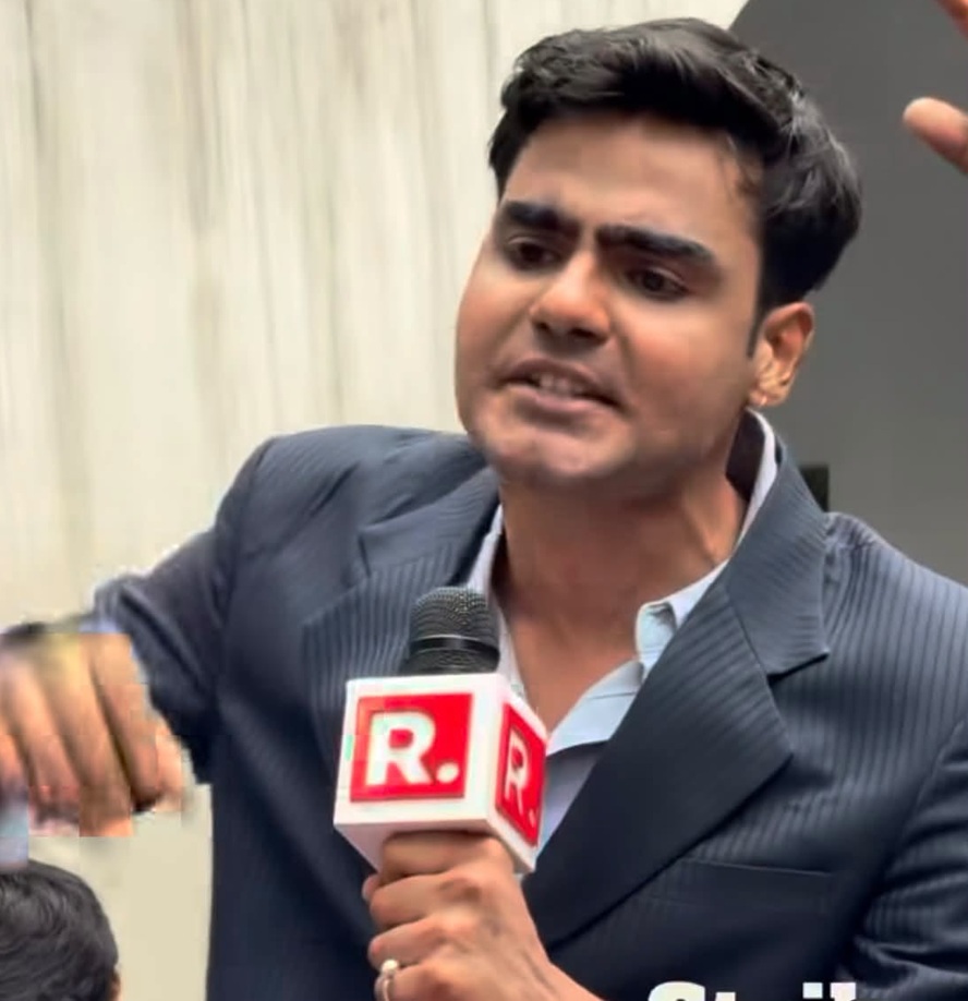 Male news reporter in a pinstripe suit speaking into a mic with a red R logo.