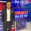 Man in a black suit and white shirt posing in a Times Now TV studio backdrop with large newsroom screens.
