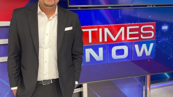 Man in a black suit and white shirt posing in a Times Now TV studio backdrop with large newsroom screens.