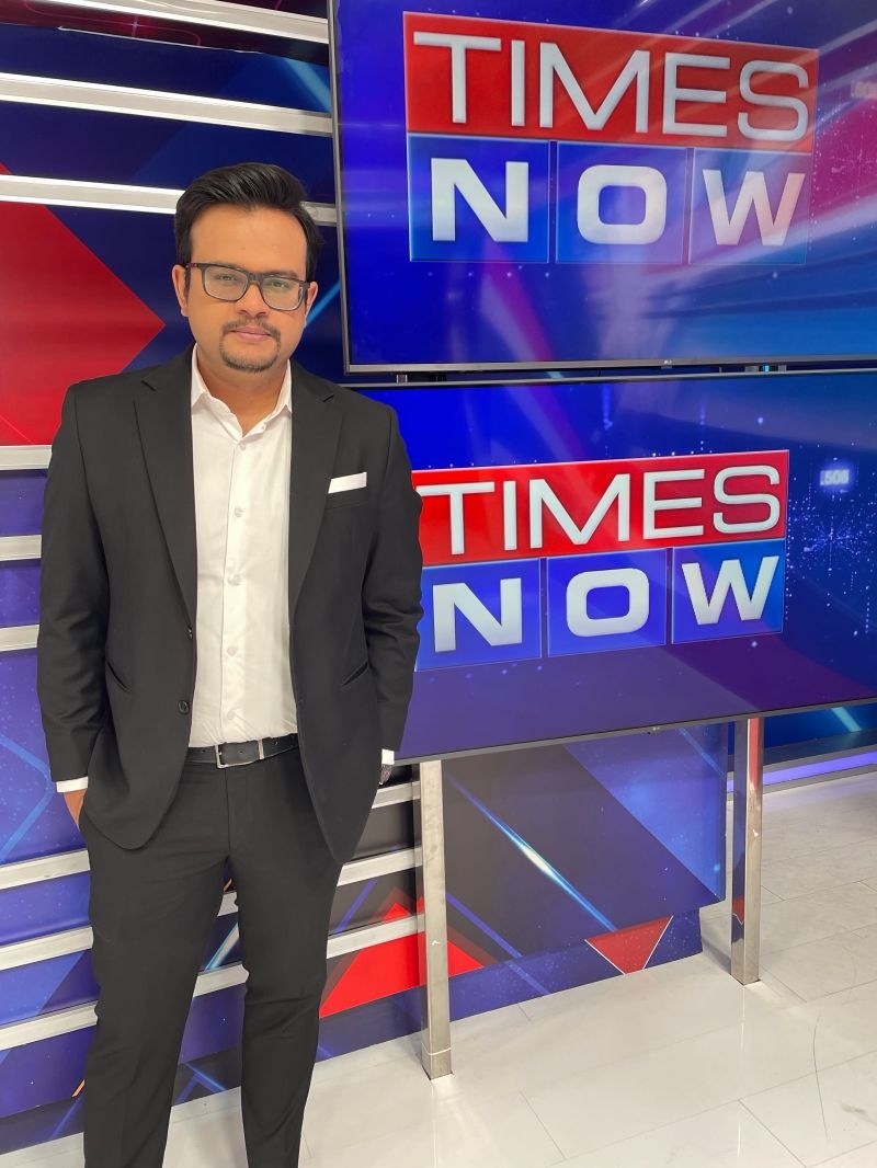Man in a black suit and white shirt posing in a Times Now TV studio backdrop with large newsroom screens.