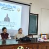 Panel of four men seated at a wooden table during a Hindi literature event; projector screen shows Hindi text and a portrait, garlanded photo on table, woman speaks at podium to the right.