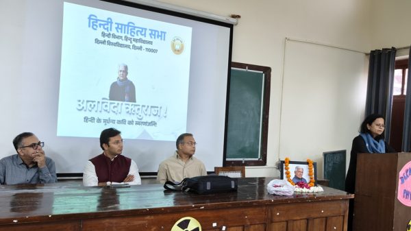 Panel of four men seated at a wooden table during a Hindi literature event; projector screen shows Hindi text and a portrait, garlanded photo on table, woman speaks at podium to the right.