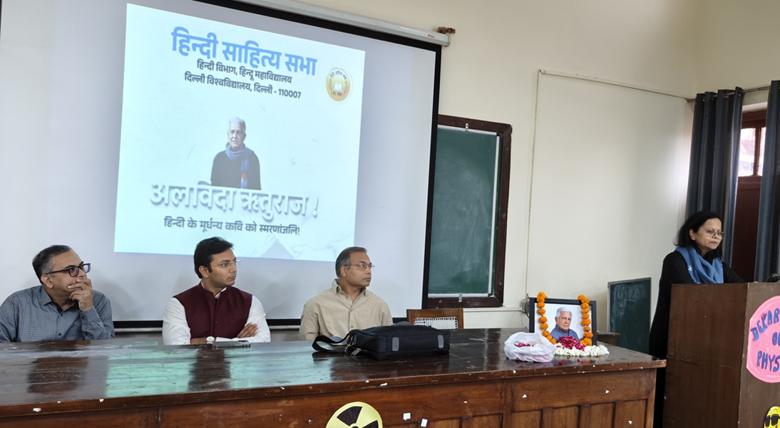 Panel of four men seated at a wooden table during a Hindi literature event; projector screen shows Hindi text and a portrait, garlanded photo on table, woman speaks at podium to the right.