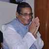 Man wearing glasses and a light blue vest, hands pressed together in a namaste gesture, tilak on the forehead.