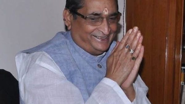 Man wearing glasses and a light blue vest, hands pressed together in a namaste gesture, tilak on the forehead.