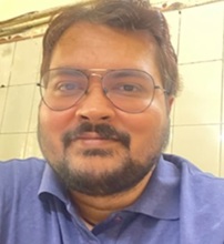 Portrait of a man with short dark hair, beard, and glasses wearing a blue polo shirt, smiling at the camera in a tiled room.