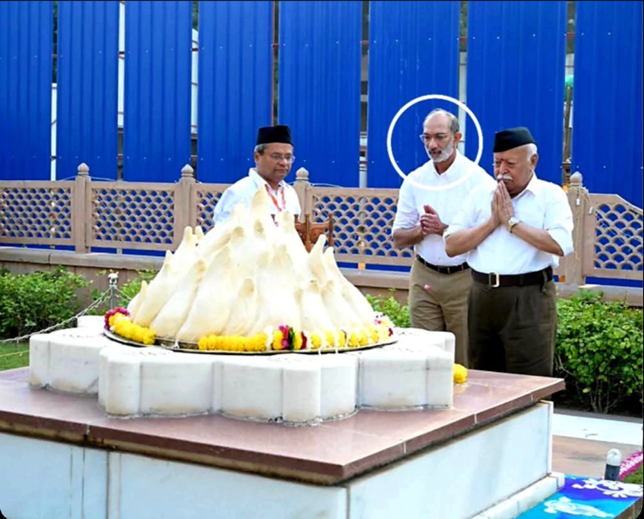 Three men in prayer beside a large white ceremonial centerpiece topped with a mound and marigold garlands at a temple-like site.