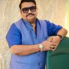 Man in a blue traditional jacket leaning on a green chair, wearing sunglasses and a watch.