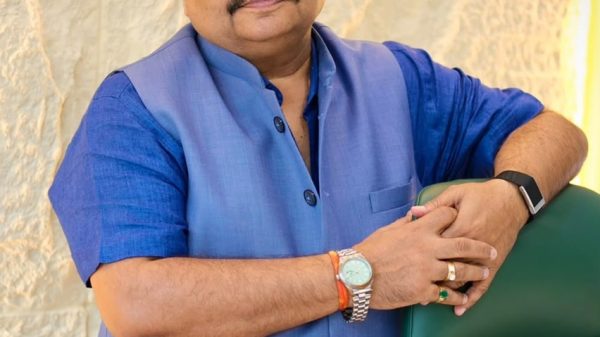 Man in a blue traditional jacket leaning on a green chair, wearing sunglasses and a watch.