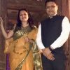 A man in a white shirt and black vest stands beside a woman in a yellow saree with green borders, posing for a photo indoors with a wooden background.