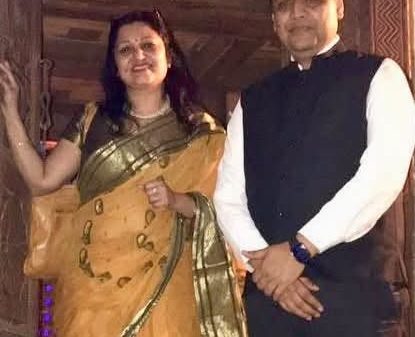 A man in a white shirt and black vest stands beside a woman in a yellow saree with green borders, posing for a photo indoors with a wooden background.