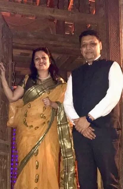 A man in a white shirt and black vest stands beside a woman in a yellow saree with green borders, posing for a photo indoors with a wooden background.