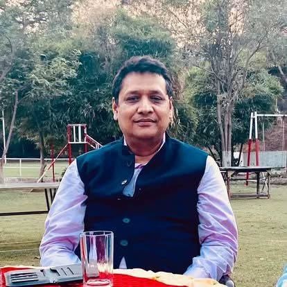 Man wearing a dark blue vest over a lavender shirt sits at a table in a park, with a glass on the table and a playground in the background.