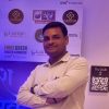 Man in a light shirt with arms crossed, posing in front of a sponsor wall featuring healthcare logos at a conference or event.