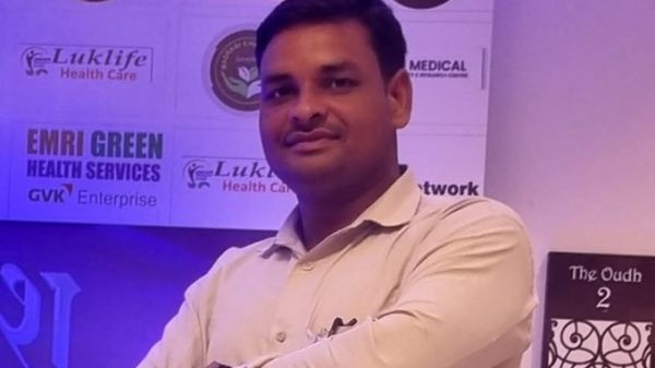 Man in a light shirt with arms crossed, posing in front of a sponsor wall featuring healthcare logos at a conference or event.
