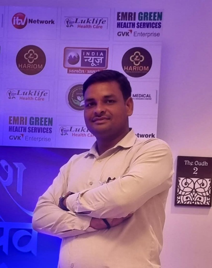 Man in a light shirt with arms crossed, posing in front of a sponsor wall featuring healthcare logos at a conference or event.
