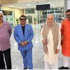 Four men stand in a bright, spacious lobby posing for a photo; one wears a lavender shirt, one in a blue suit, one in a beige vest and kurta, and another in an orange kurta; they smile subtly at the camera.
