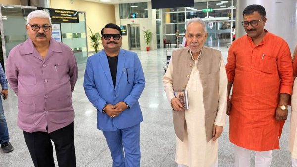 Four men stand in a bright, spacious lobby posing for a photo; one wears a lavender shirt, one in a blue suit, one in a beige vest and kurta, and another in an orange kurta; they smile subtly at the camera.