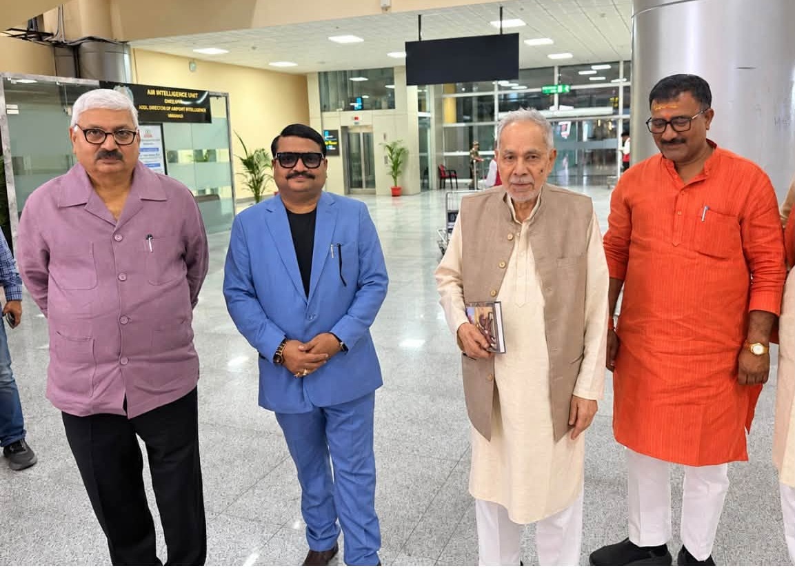 Four men stand in a bright, spacious lobby posing for a photo; one wears a lavender shirt, one in a blue suit, one in a beige vest and kurta, and another in an orange kurta; they smile subtly at the camera.