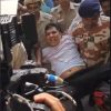 Police officers restrain a man in a white shirt on a car hood as a crowd films the scene with cameras nearby.