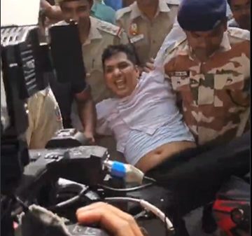 Police officers restrain a man in a white shirt on a car hood as a crowd films the scene with cameras nearby.