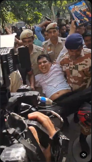 Police officers restrain a man in a white shirt on a car hood as a crowd films the scene with cameras nearby.