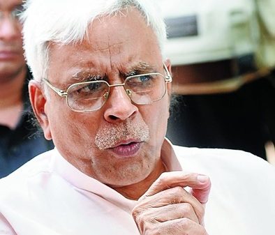 Senior man with white hair and glasses, in a pale pink shirt, gesturing with his hand while speaking.