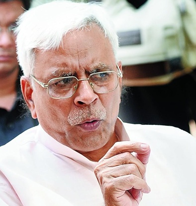 Senior man with white hair and glasses, in a pale pink shirt, gesturing with his hand while speaking.