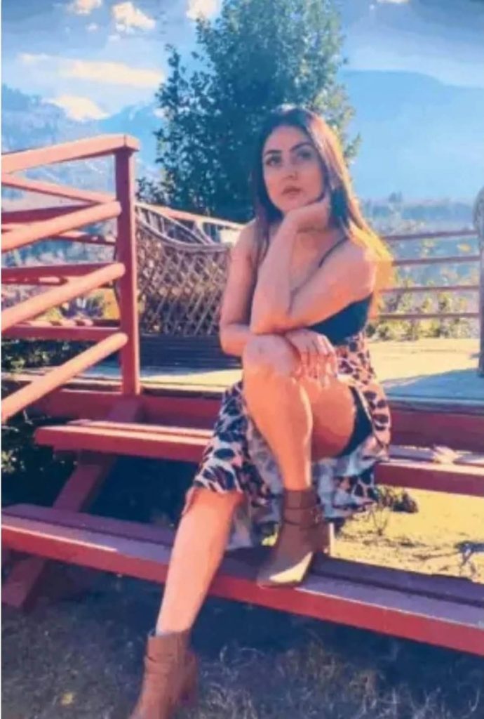 Woman sitting on a red wooden bench outdoors, resting chin on her hand, wearing a patterned dress and brown ankle boots with a black top; mountains in the background.