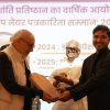 An elderly man with glasses hands a wooden plaque to a smiling man on stage, with another man watching and a Hindi banner in the background on stage.