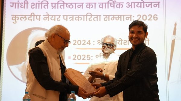 An elderly man with glasses hands a wooden plaque to a smiling man on stage, with another man watching and a Hindi banner in the background on stage.