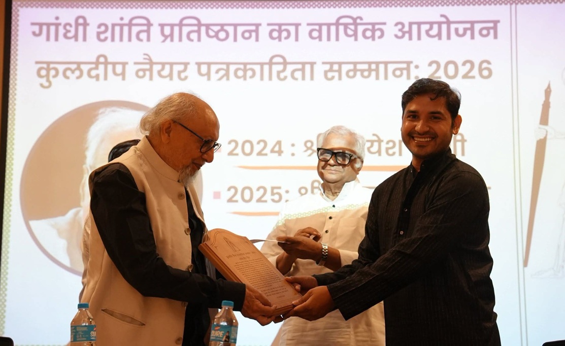 shyam_2 | No. 1 Indian Media News Portal An elderly man with glasses hands a wooden plaque to a smiling man on stage, with another man watching and a Hindi banner in the background on stage.
