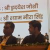 Two men seated on a stage at a panel discussion, with water bottles and marigold garlands on the table and a Hindi slide in the background.