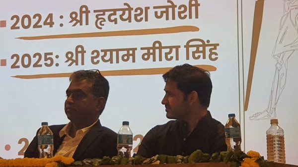 Two men seated on a stage at a panel discussion, with water bottles and marigold garlands on the table and a Hindi slide in the background.