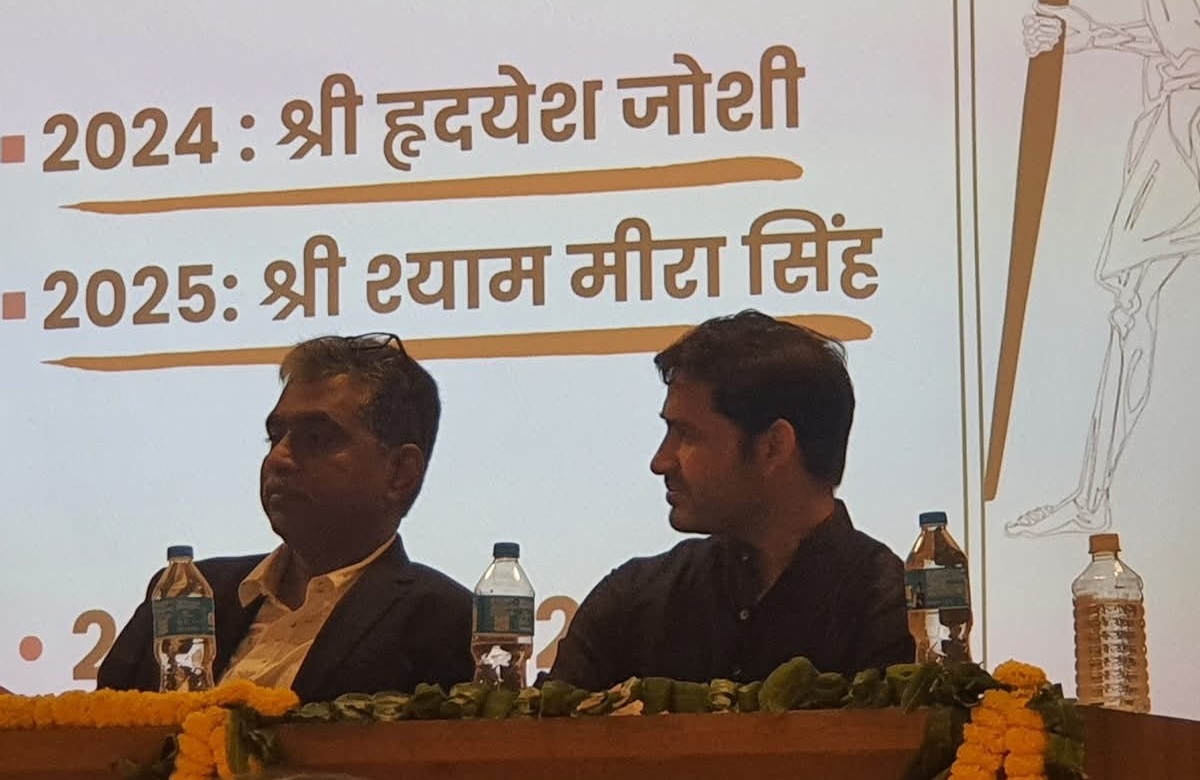shyam_hradyesh | No. 1 Indian Media News Portal Two men seated on a stage at a panel discussion, with water bottles and marigold garlands on the table and a Hindi slide in the background.