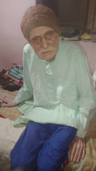 Elderly person wearing a light green kurta and brown headscarf sits on a bed in a softly lit room, looking at the camera.