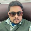 Man with beard and mustache wearing sunglasses and a green vest, seated against a dark brown headboard? (portrait)
