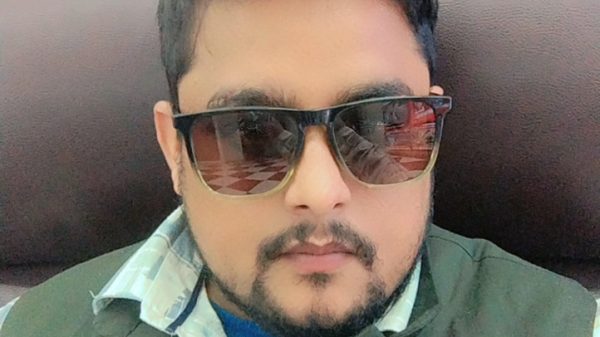 Man with beard and mustache wearing sunglasses and a green vest, seated against a dark brown headboard? (portrait)