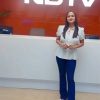 Woman in a white blouse and blue pants stands at a white reception desk in front of a red NDTV sign on the wall behind her.