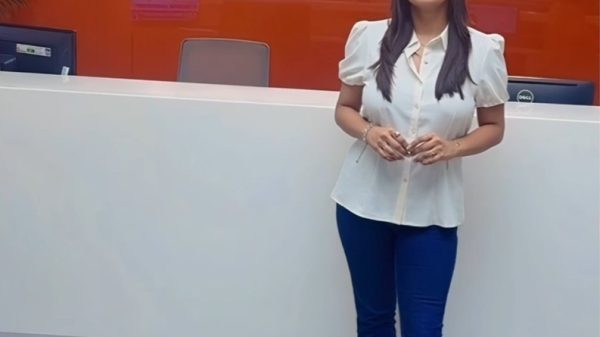 Woman in a white blouse and blue pants stands at a white reception desk in front of a red NDTV sign on the wall behind her.