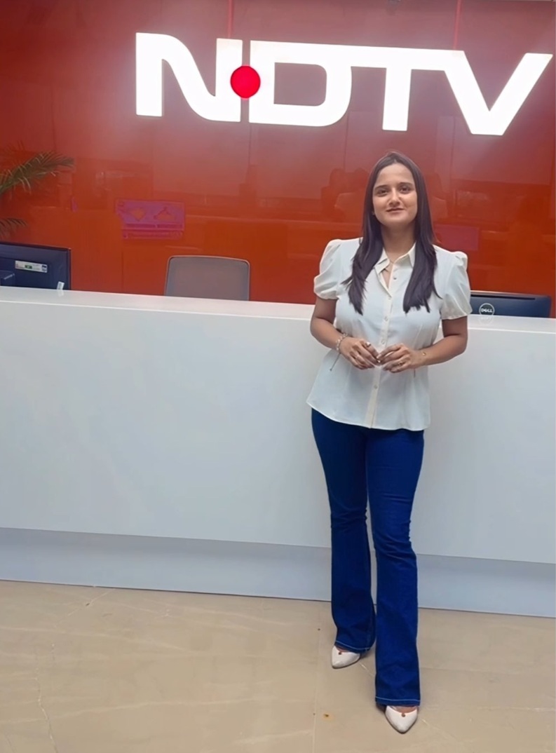 Woman in a white blouse and blue pants stands at a white reception desk in front of a red NDTV sign on the wall behind her.
