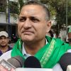 Man wearing a green scarf speaks to reporters, microphones in front and a crowd behind him in an outdoor setting.