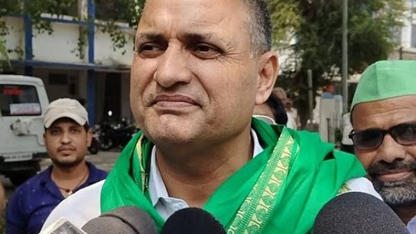 Man wearing a green scarf speaks to reporters, microphones in front and a crowd behind him in an outdoor setting.