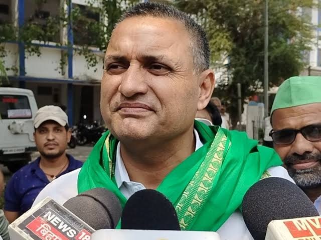 Man wearing a green scarf speaks to reporters, microphones in front and a crowd behind him in an outdoor setting.