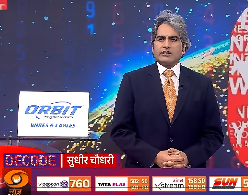 Male news anchor in a dark suit and orange tie stands in a TV studio, with an Orbit Wires & Cables sign on a lectern to his left and a colorful digital backdrop behind him.