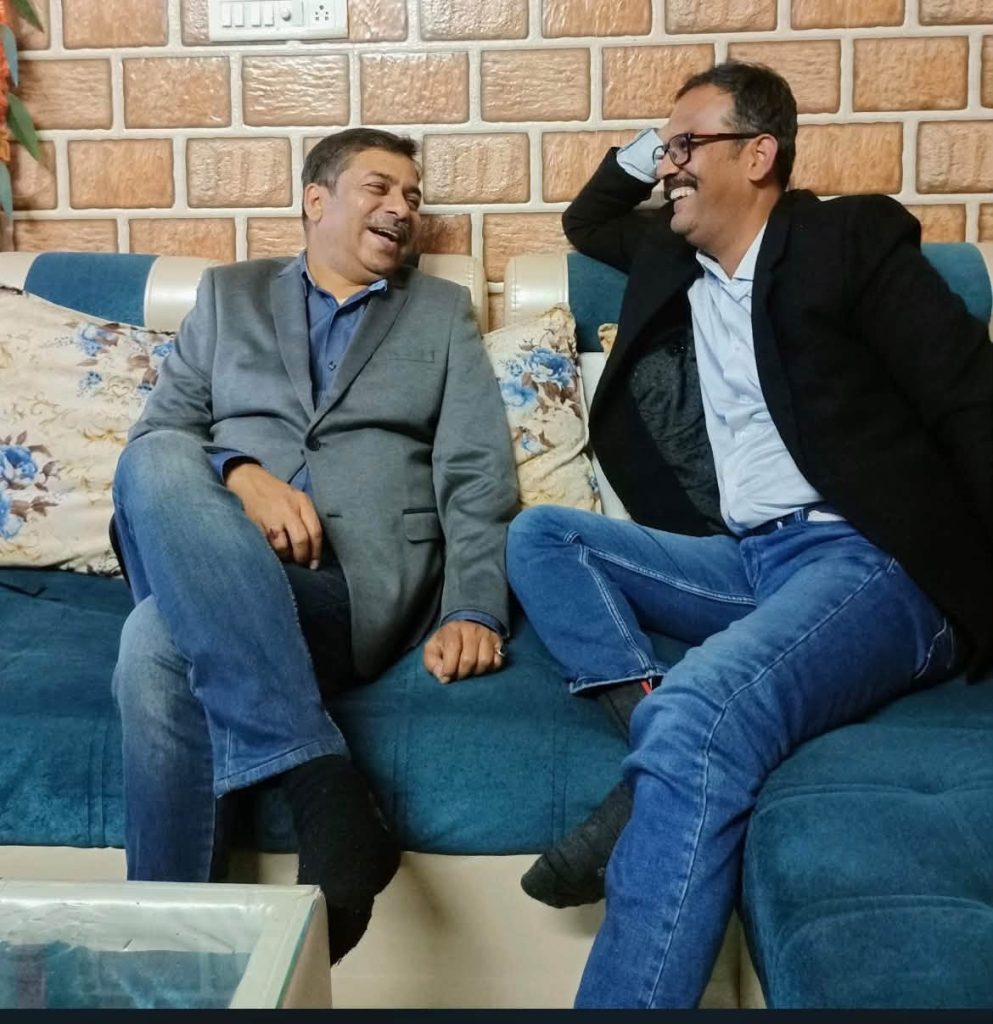 Two men sit on a blue sofa, smiling and laughing together in a casual living room setting.