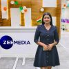 Woman in a navy dress stands at a Zee Media reception desk with Indian flag pin and festive balloons in the background.