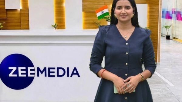 Woman in a navy dress stands at a Zee Media reception desk with Indian flag pin and festive balloons in the background.