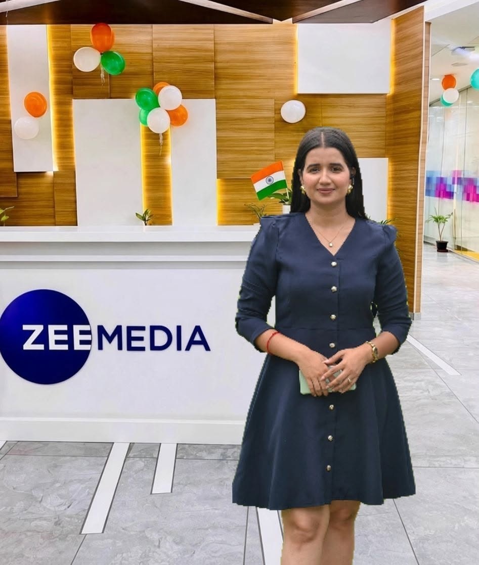 Woman in a navy dress stands at a Zee Media reception desk with Indian flag pin and festive balloons in the background.