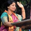 Woman in a colorful sari raising her right hand while speaking at a podium, gesture of greeting or pledge.